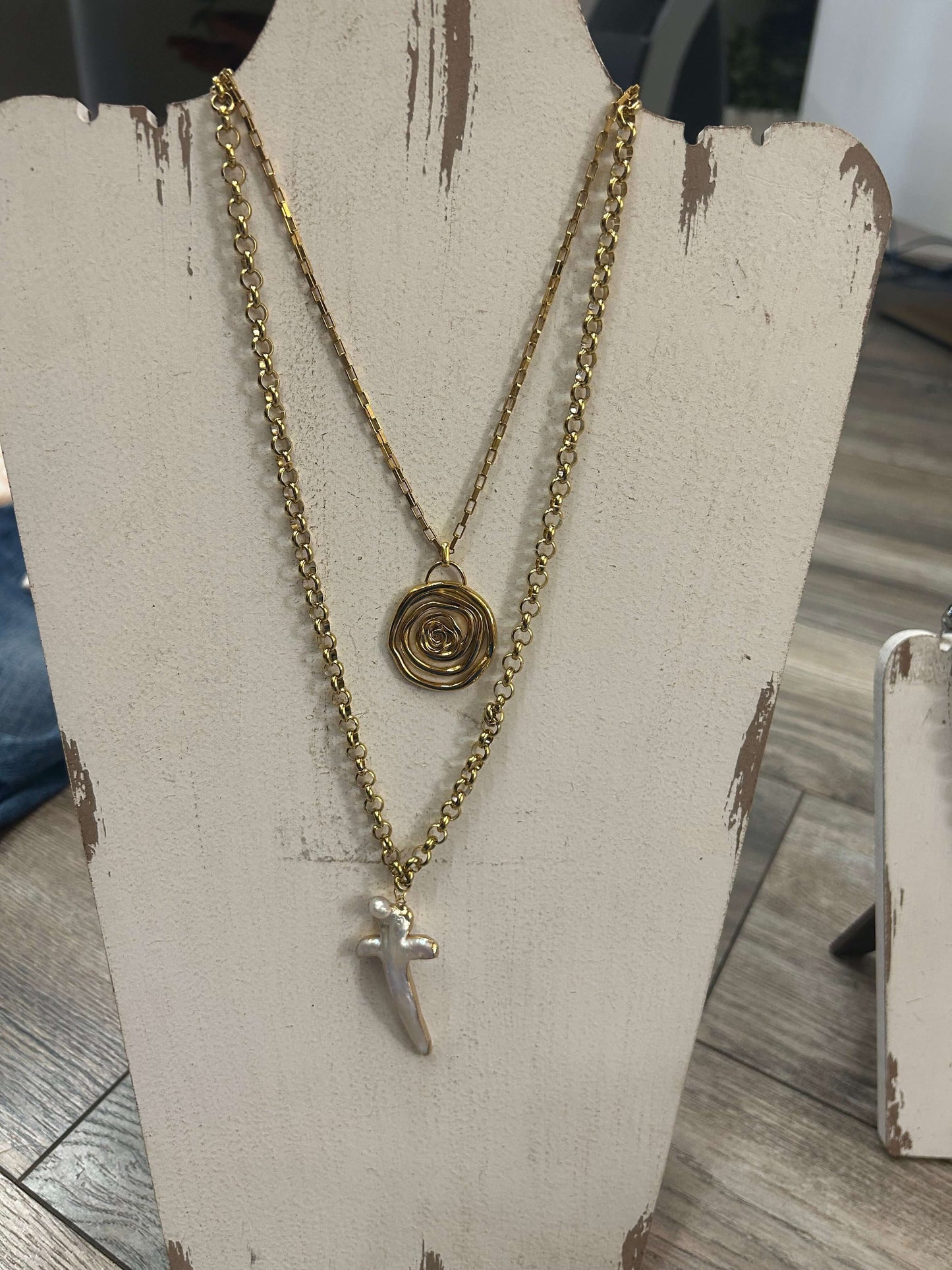 Layered gold necklace with swirly rose pendant and charm, displayed on a wooden stand.