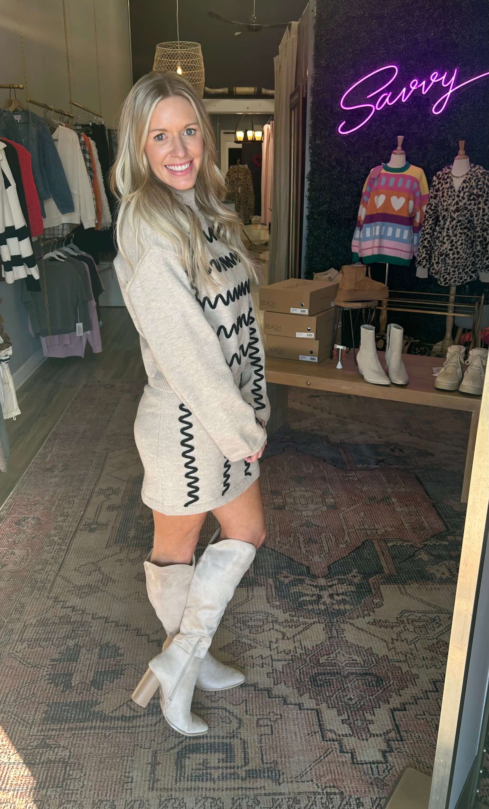Woman modeling a stylish sweater dress with wavy trim, paired with knee-high boots, in a chic boutique setting.
