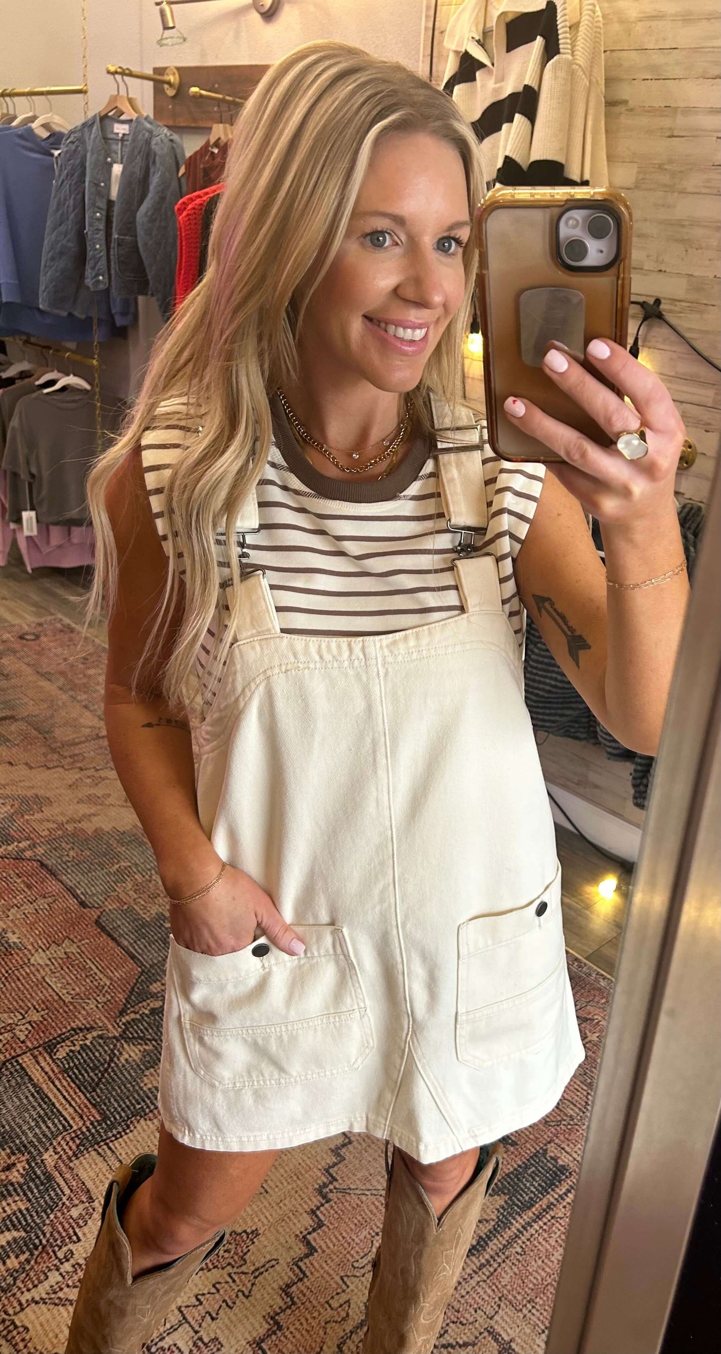 Woman in a striped shirt wearing a denim mineral washed overall dress in bone color, featuring adjustable straps and pockets.