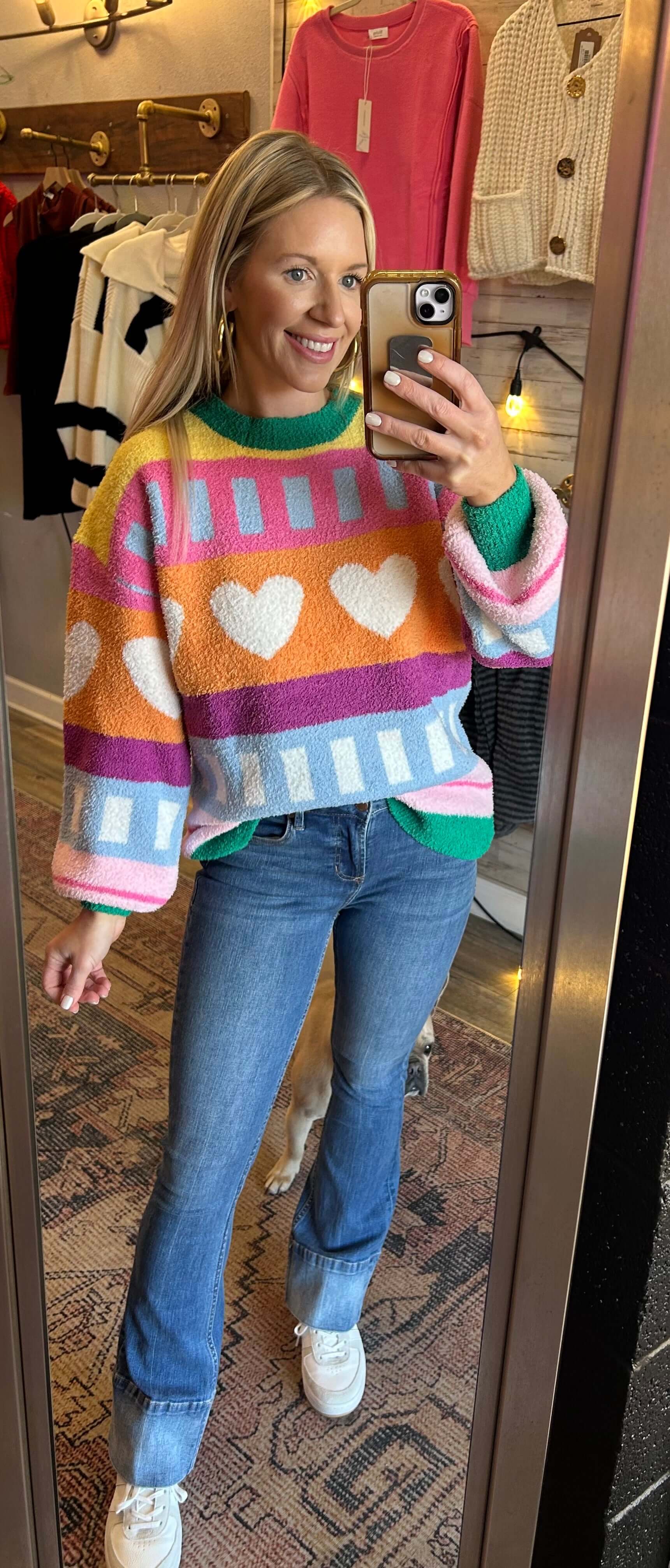 Woman in a colorful, patterned hearts knit sweater, showcasing playful winter style with jeans in a mirror.