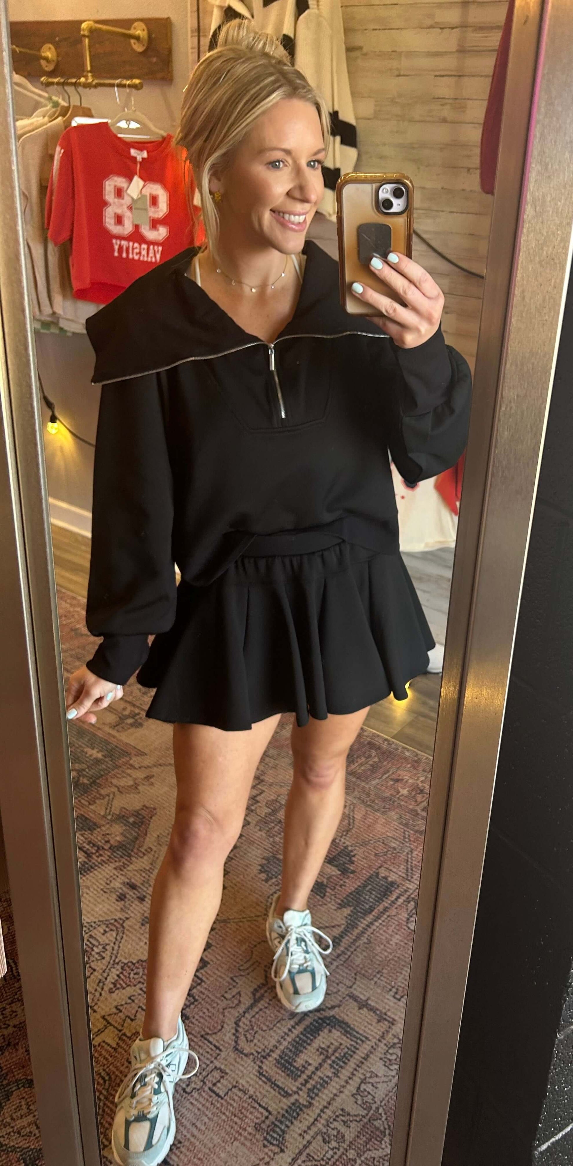 Woman in a black neoprene flare skort and pullover set with cowl turtleneck and half zip, posing in a mirror.