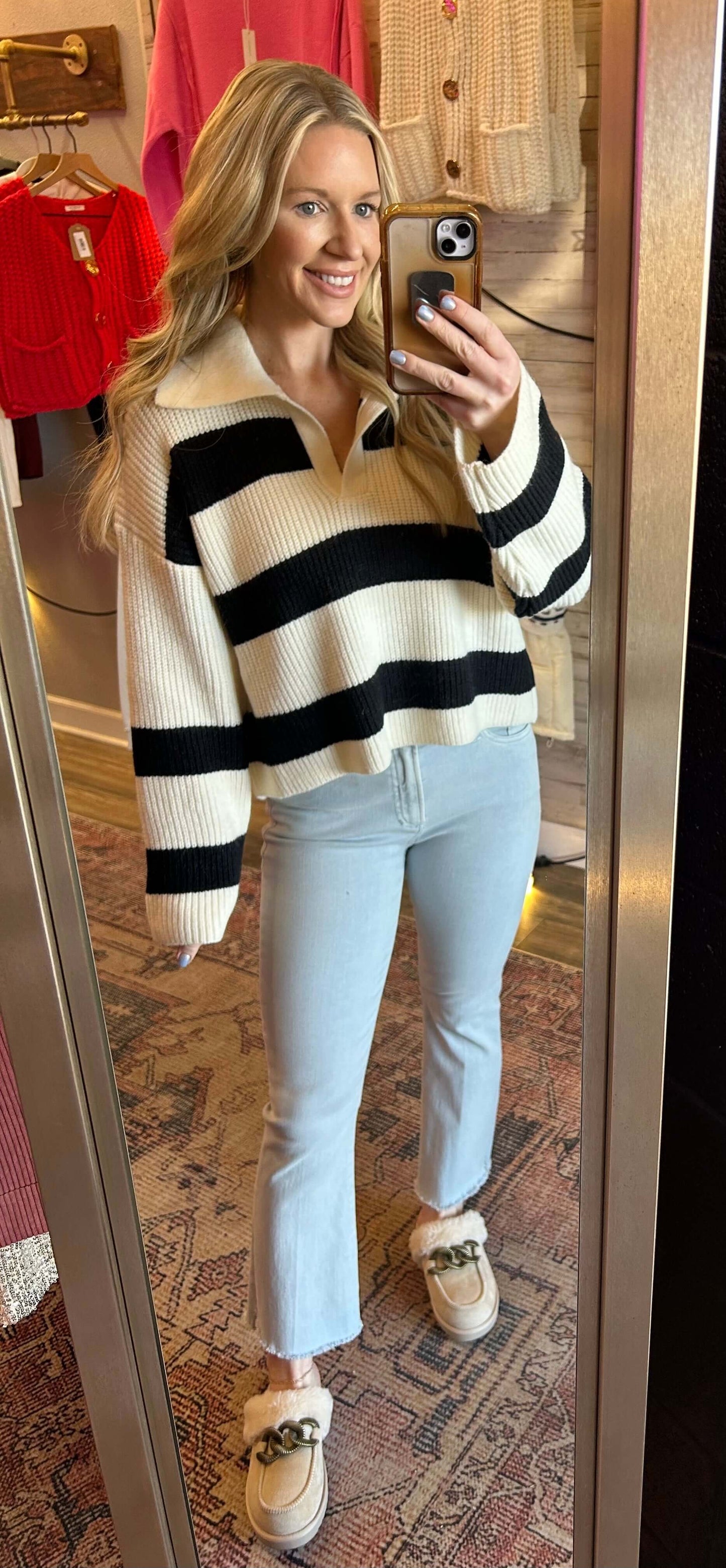 Woman wearing an ivory and black wide stripe polo knit sweater, smiling in front of a mirror, styled with light blue jeans.
